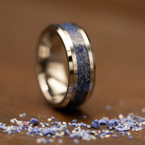 A polished silver-toned ring with a blue and white speckled inlay, standing on a wooden surface with small blue and white stones scattered in the foreground.