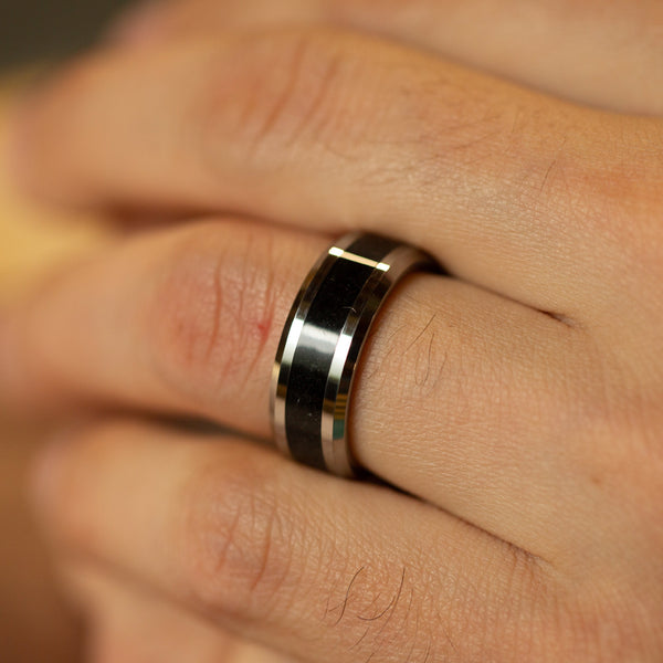A close-up of a polished silver band ring with a black inlay, worn on a person's finger.