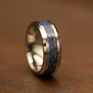 A polished silver-toned ring with a textured blue and gold speckled inlay, resting on a warm brown wooden surface.