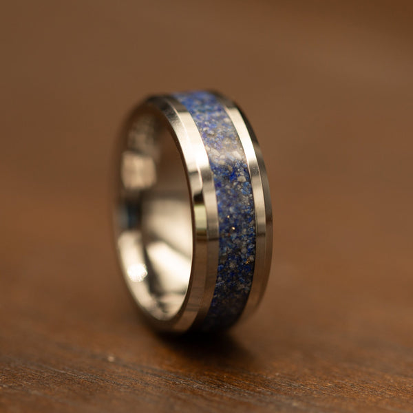 A polished silver-toned ring with a textured blue and gold speckled inlay, resting on a warm brown wooden surface.