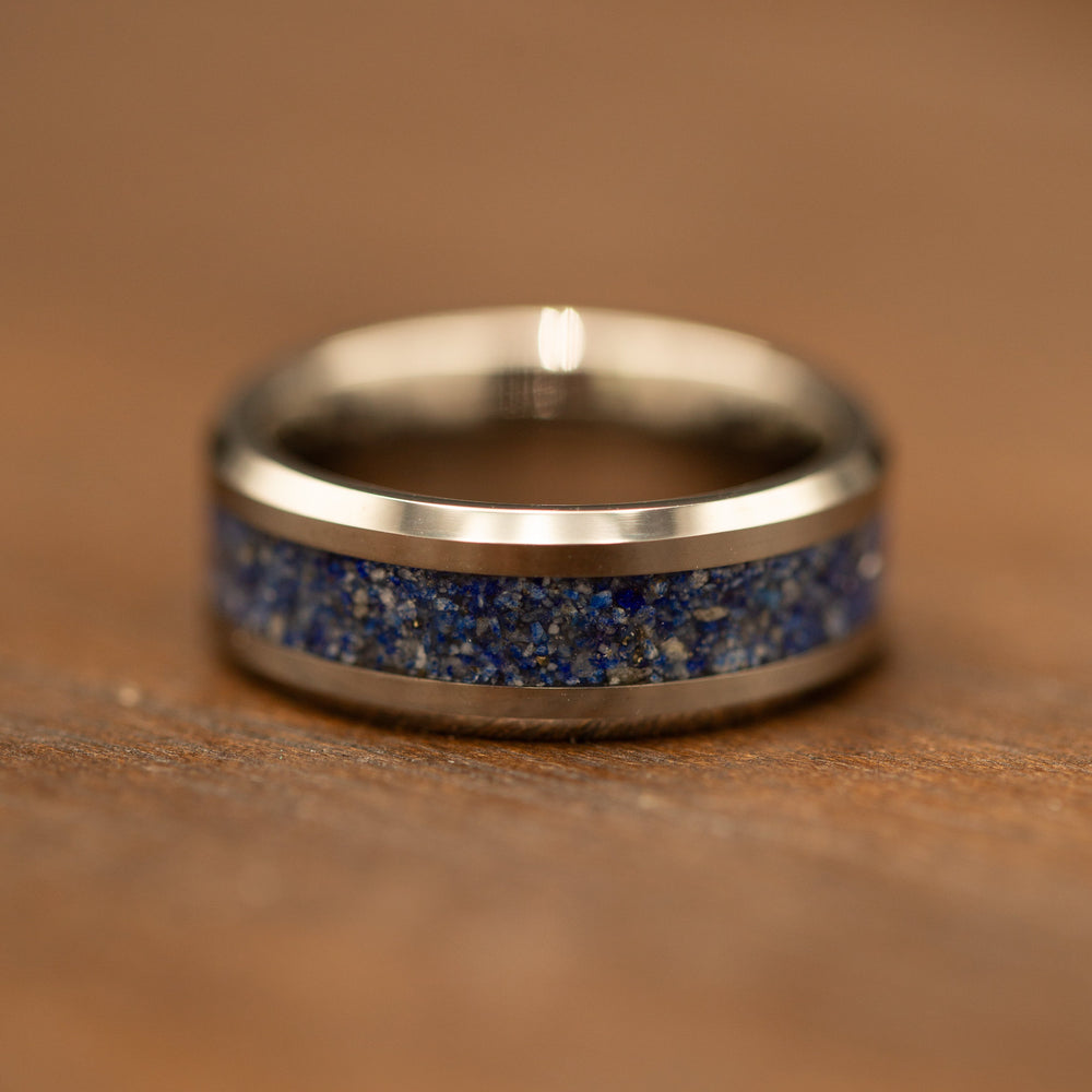 A polished silver-toned ring with a textured blue and gold speckled inlay, resting on a warm brown wooden surface.